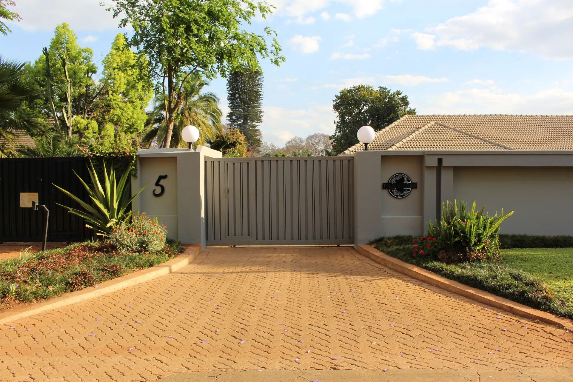 Paved driveway leading to the gated entrance of FIRN Lodge in Belvedere, Harare, with globe lamps, the FIRN Lodge sign, flowering beds and palm trees under a blue sky